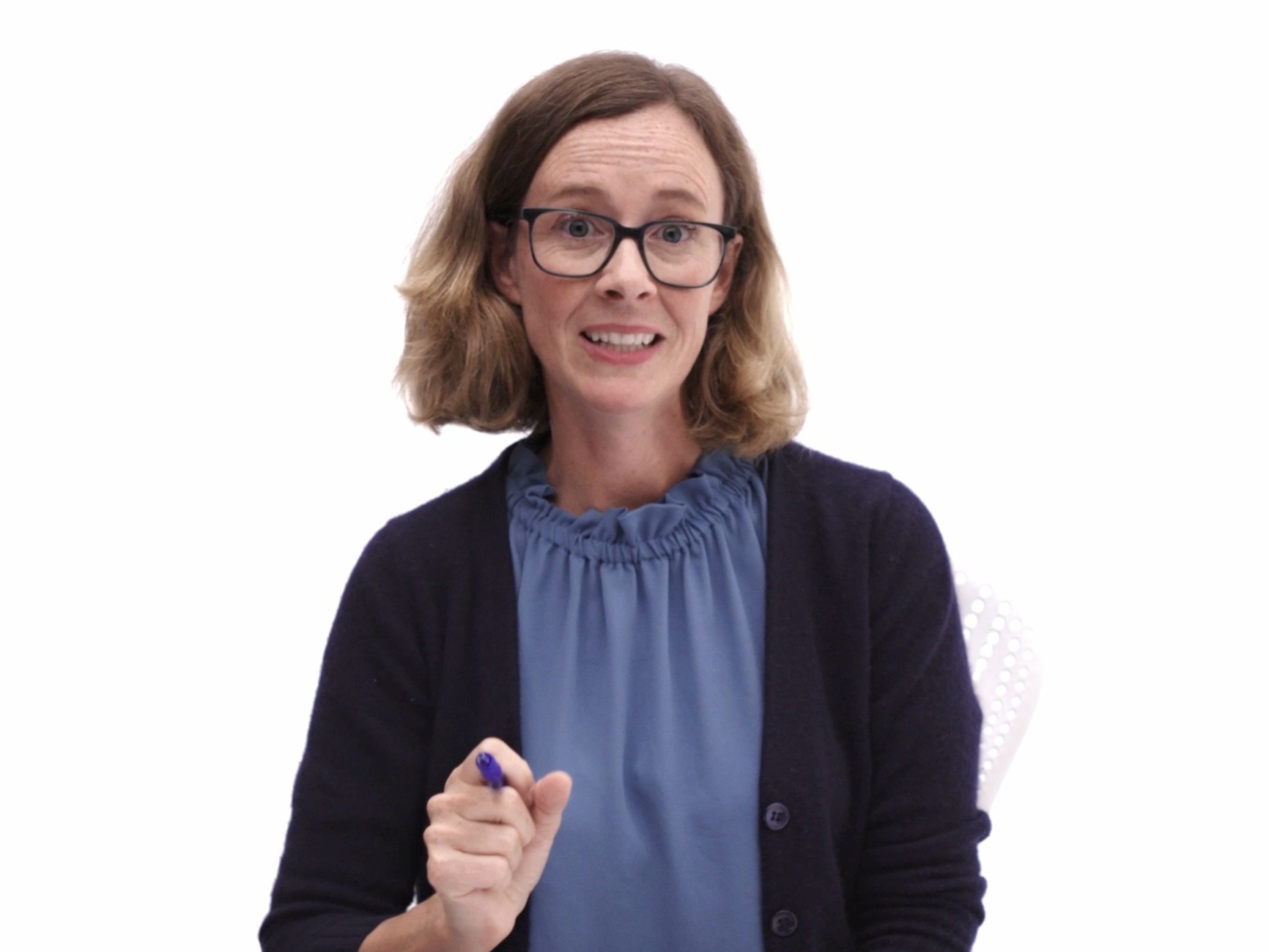 A white woman with medium-length red-blonde hair and large blue glasses talks in front of a white background gesturing with a pen as she wears a dark blue cardigan over a light blue blouse.