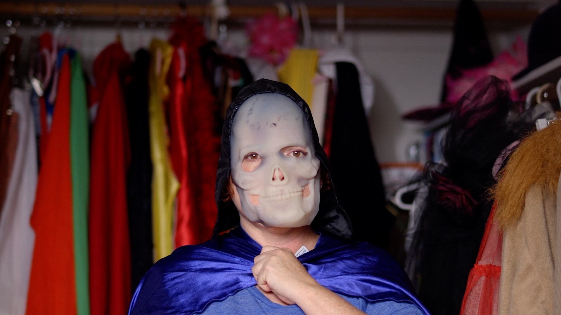 A woman wearing a skull mask and a bright blue cape stands in a closet full of costumes.