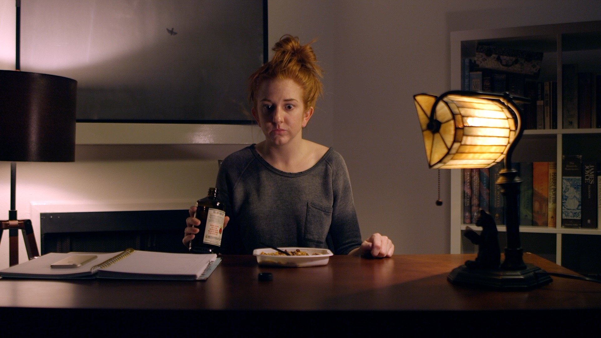 A redhaired woman with her head in a messy bun sits at a desk staring blankly ahead.