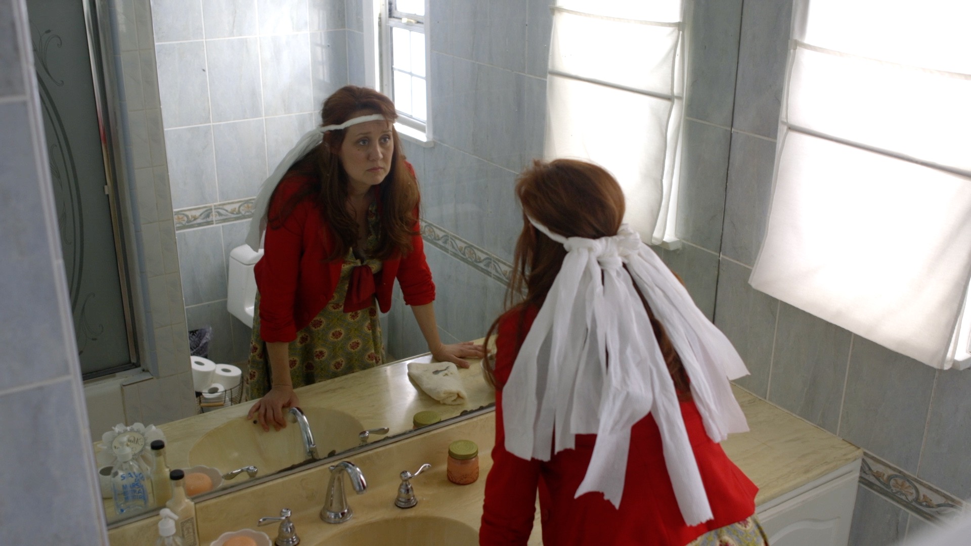 A woman in a dress wearing a tissue paper headband stares wide-eyed into a bathroom mirror.