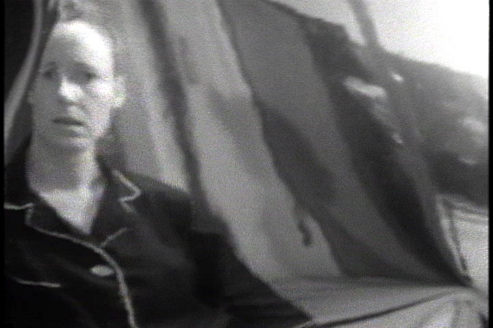 Black and white photo of a white woman in dark pajamas staring into a warped bathroom mirror.