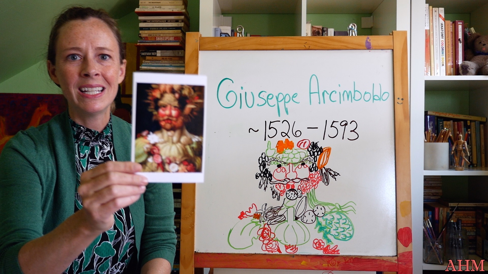 smiling Joan Jill wears a green cardigan and dress and holds up a photo of an Archimbaldo painting while sitting next to a whiteboard with the artist's name on it
